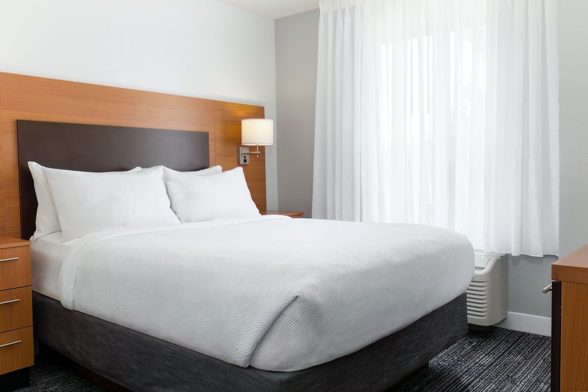 A neatly made bed with white linens, a wooden headboard, a bedside table with a lamp, and a window with sheer curtains.