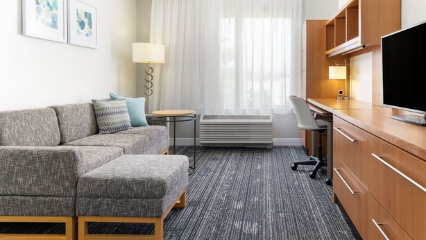 Une chambre d’hôtel avec un canapé gris, un bureau en bois, une télévision et une fenêtre avec des rideaux blancs.