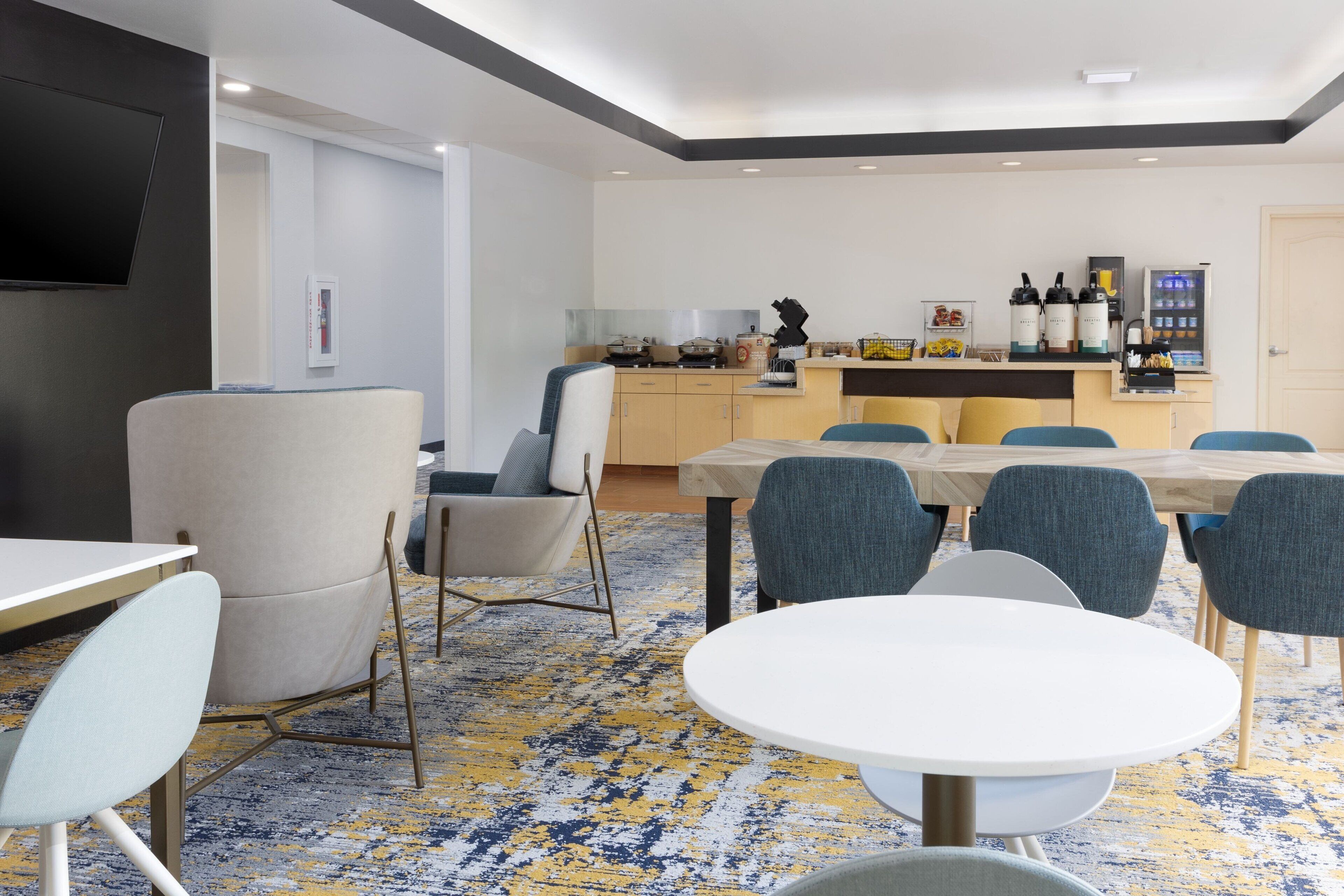 A modern hotel room with a dining area, a kitchen with appliances, and a TV mounted on the wall.