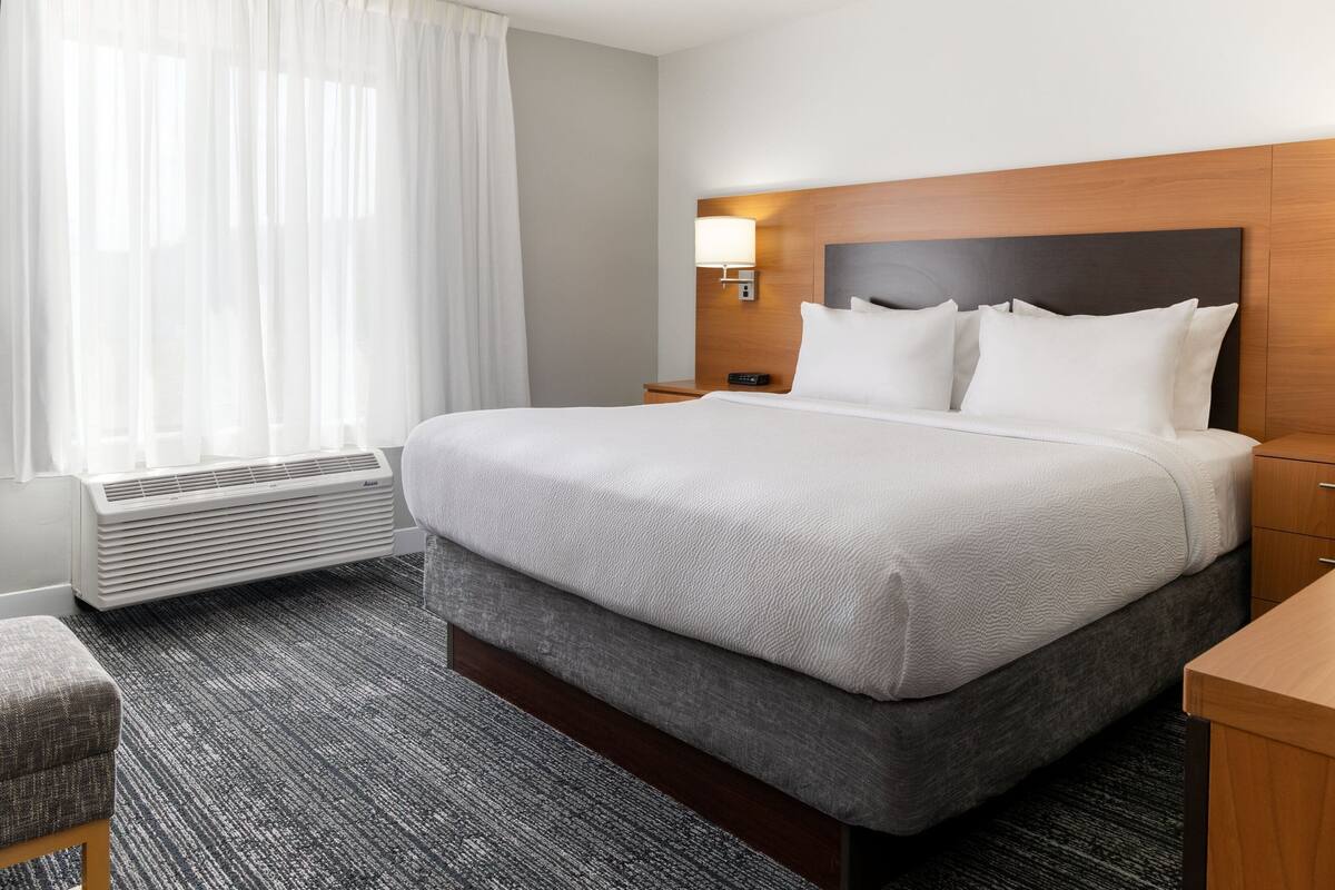 A hotel room with a large bed, a bedside table, a lamp, a window with curtains, and a radiator.