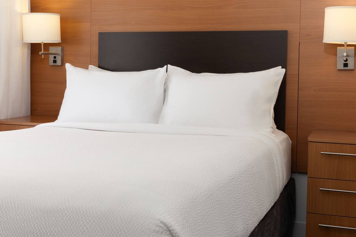 A neatly made bed with white bedding and a dark headboard, flanked by wall-mounted lamps and a wooden nightstand.