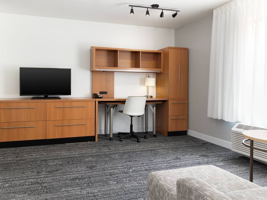 A modern hotel room with a wooden desk, a flat-screen TV, a white office chair, a small table, and a large window with white curtains.