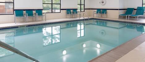 Una piscina interior con agua cristalina, rodeada de sillas reclinables color turquesa y un suelo de baldosas.