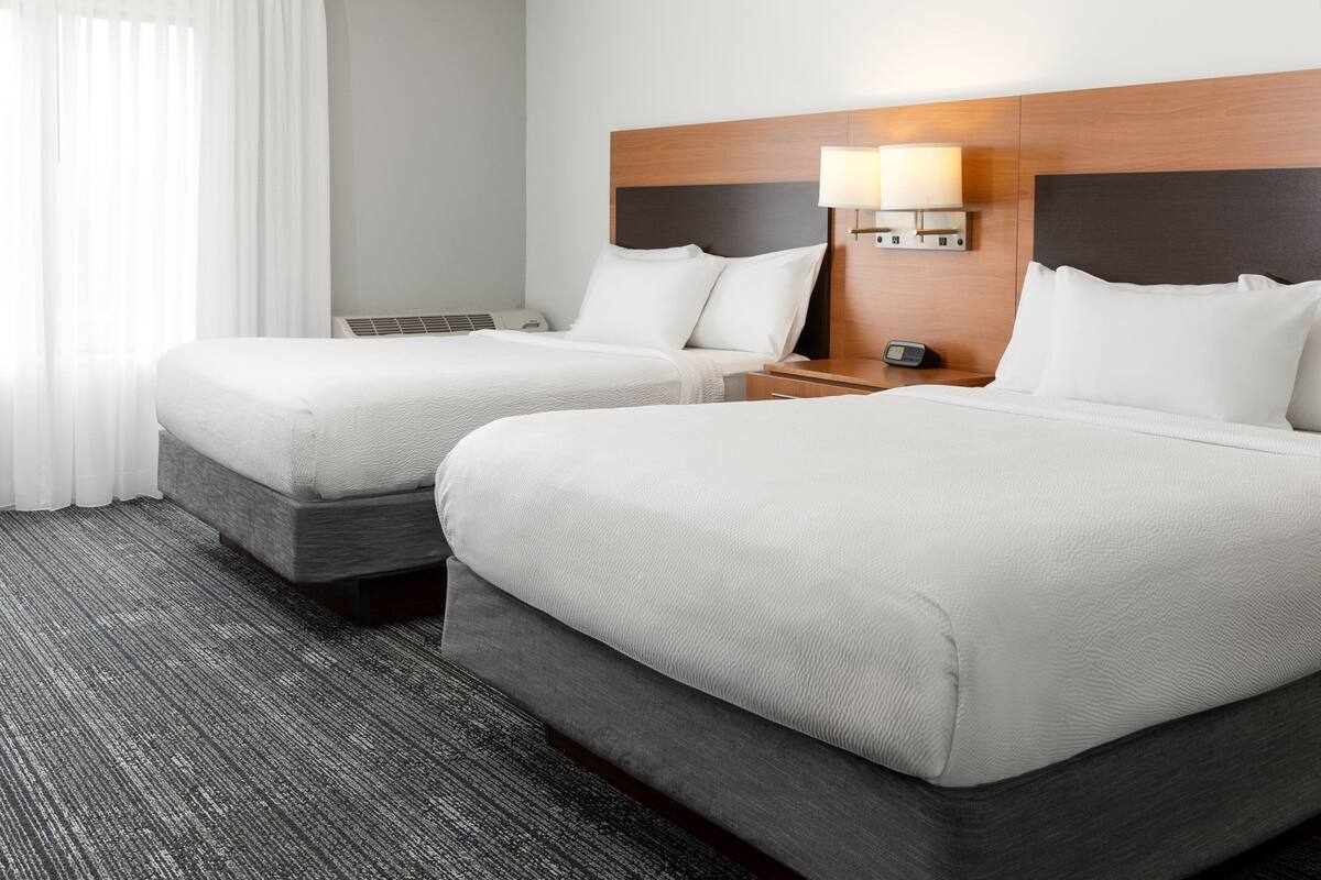 A hotel room with two beds, a wooden headboard, a bedside table with a lamp, and a window with white curtains.