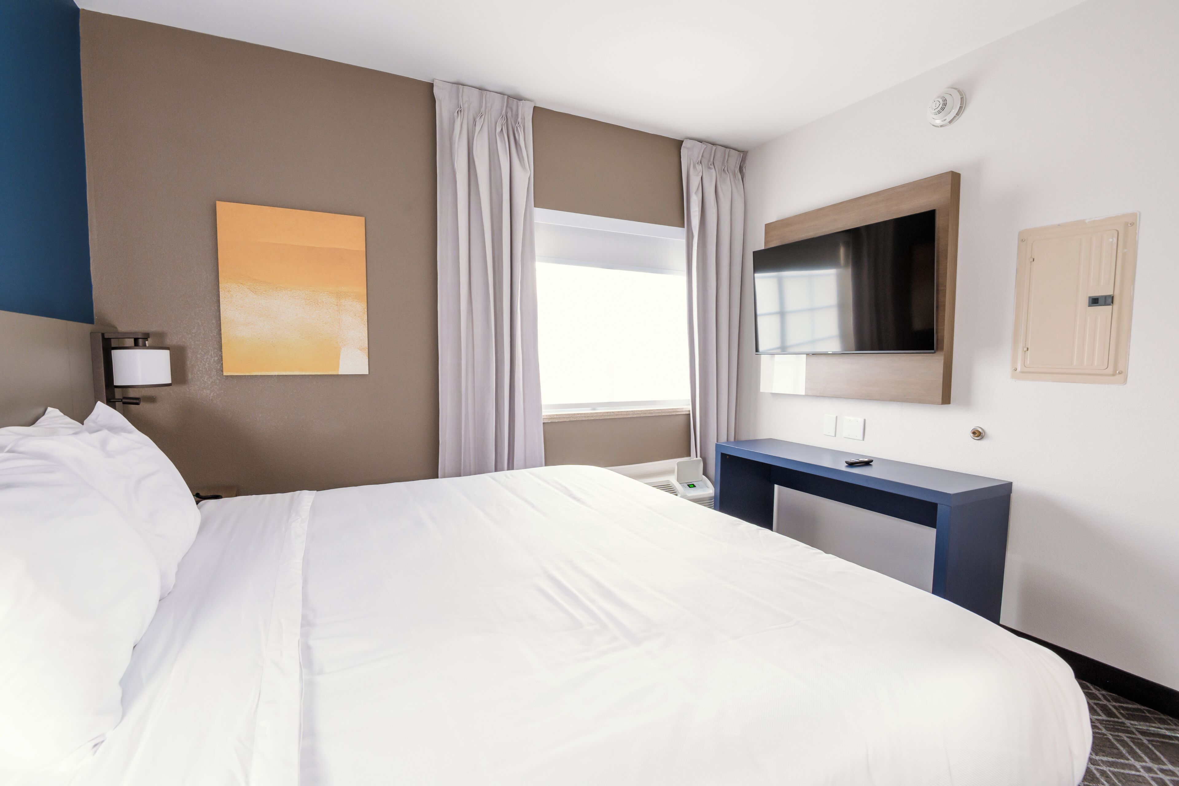 A hotel room with a large bed, a desk, a flat-screen TV, a window with curtains, and a wall-mounted artwork.