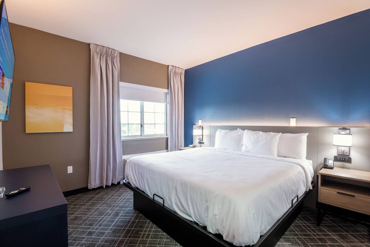 A hotel room with a bed, bedside table, lamp, and a window with curtains.