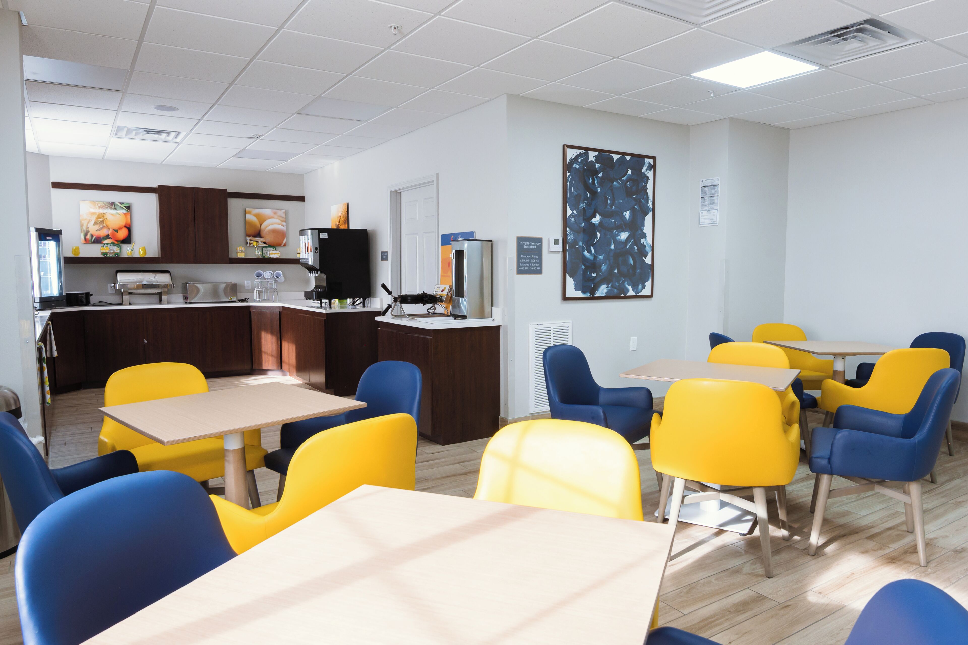 Un coin repas avec des chaises jaunes et bleues, une cuisine à l’arrière-plan et un grand tableau abstrait accroché au mur.