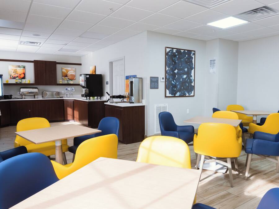 Un coin repas avec des chaises jaunes et bleues, une cuisine à l’arrière-plan et un grand tableau abstrait accroché au mur.