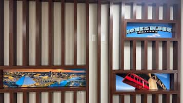 Pared com ripas verticais de madeira e duas molduras. Uma moldura mostra uma paisagem urbana com uma ponte e um rio, e a outra apresenta um trem vermelho sobre os trilhos.