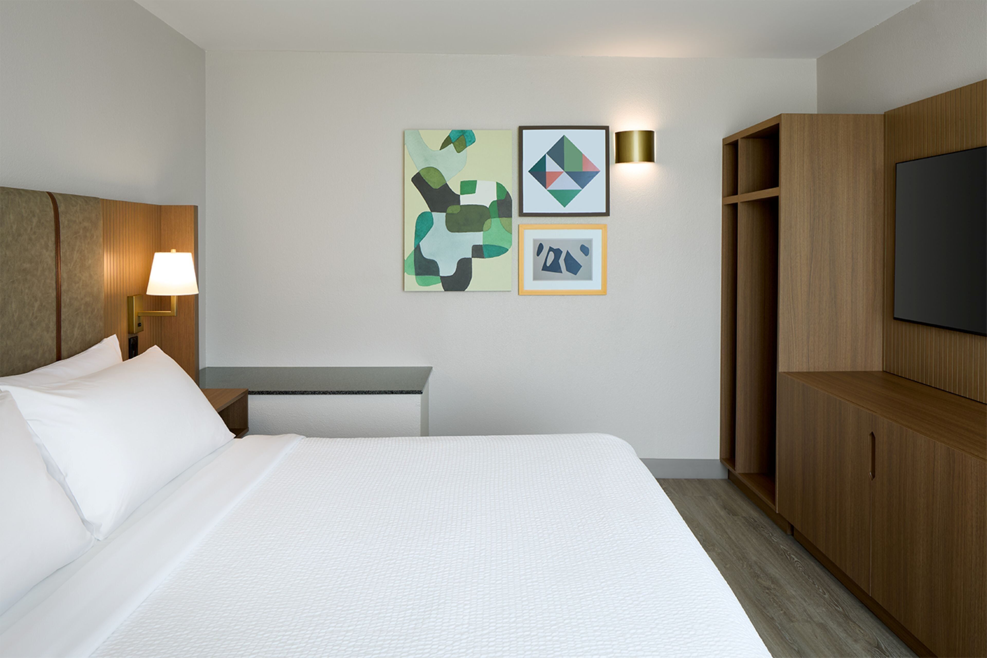 A hotel room with a bed, a desk, a lamp, a TV, and artwork on the wall.