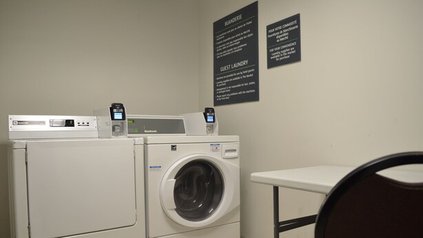 Un área de lavandería con una lavadora de carga frontal, una secadora y un panel de control. Se observan carteles que indican servicios de lavandería para huéspedes e instrucciones de uso.