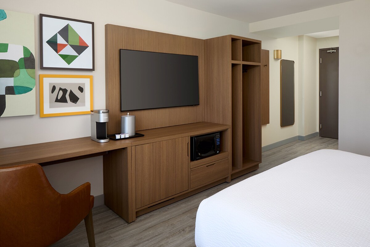 Une chambre d’hôtel équipée d’un lit, d’un téléviseur fixé à un meuble en bois, d’un four à micro-ondes, d’une machine à café et d’œuvres d’art accrochées au mur.