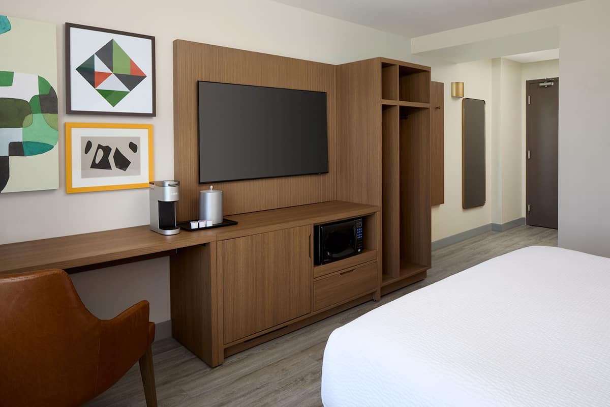 Habitación de hotel con cama, televisor montado en un mueble de madera, microondas, cafetera y una obra de arte en la pared.