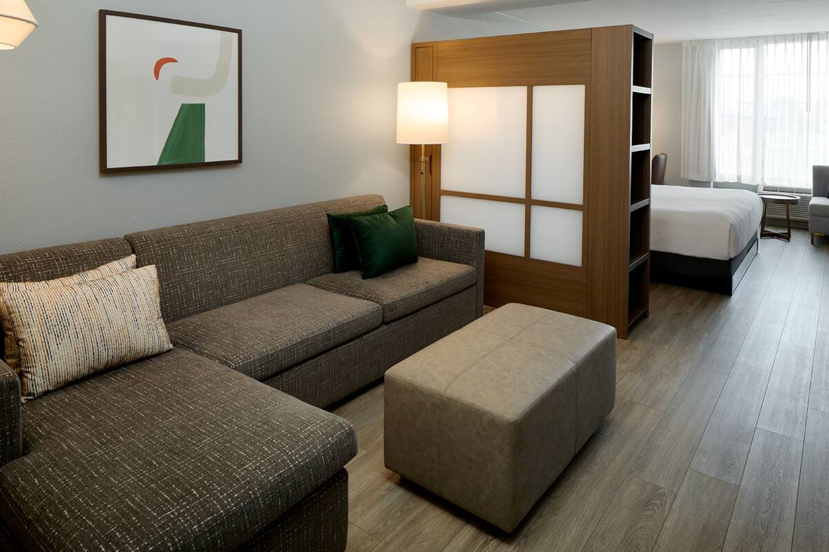 Habitación de hotel con sofá, reposapiés, cama, mesita de noche, lámpara y ventana con cortinas.
