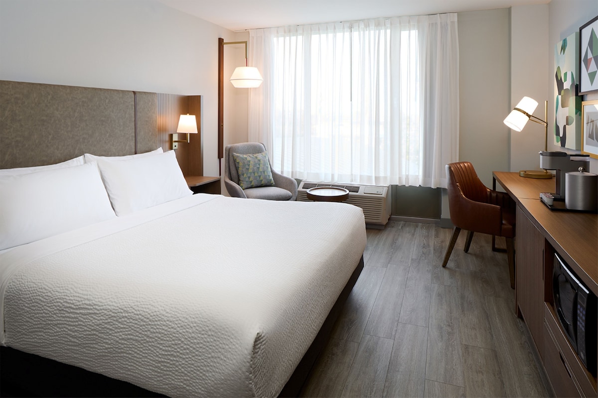 A hotel room with a large bed, a desk with a chair, a small table, and a window with curtains.