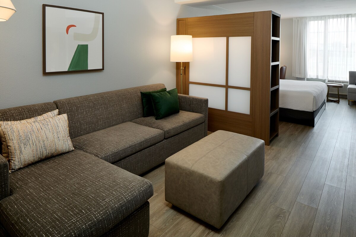 Une chambre d’hôtel comprenant un canapé, un pouf, un lit, une table de chevet, une lampe et une fenêtre avec des rideaux.
