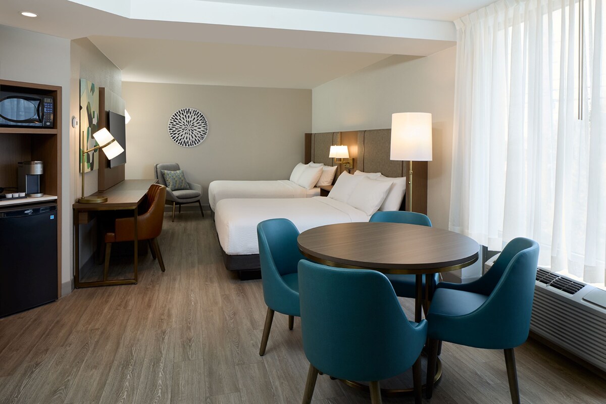 Une chambre d’hôtel comprenant un lit, deux tables de chevet, un bureau avec une chaise, une table de salle à manger ronde avec des chaises, un four à micro-ondes, une télévision et un élément décoratif mural.