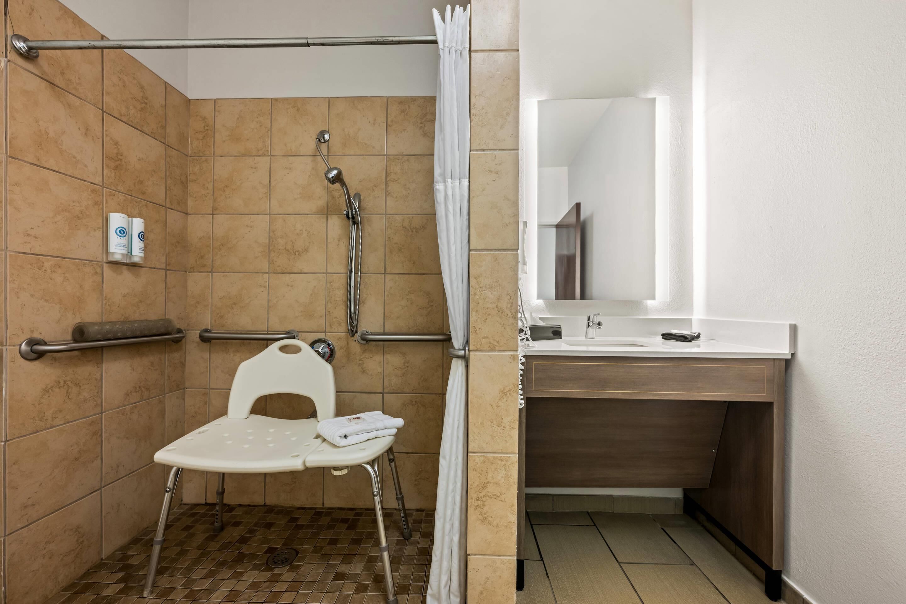 A bathroom with a shower, a white chair, a sink, and a mirror.