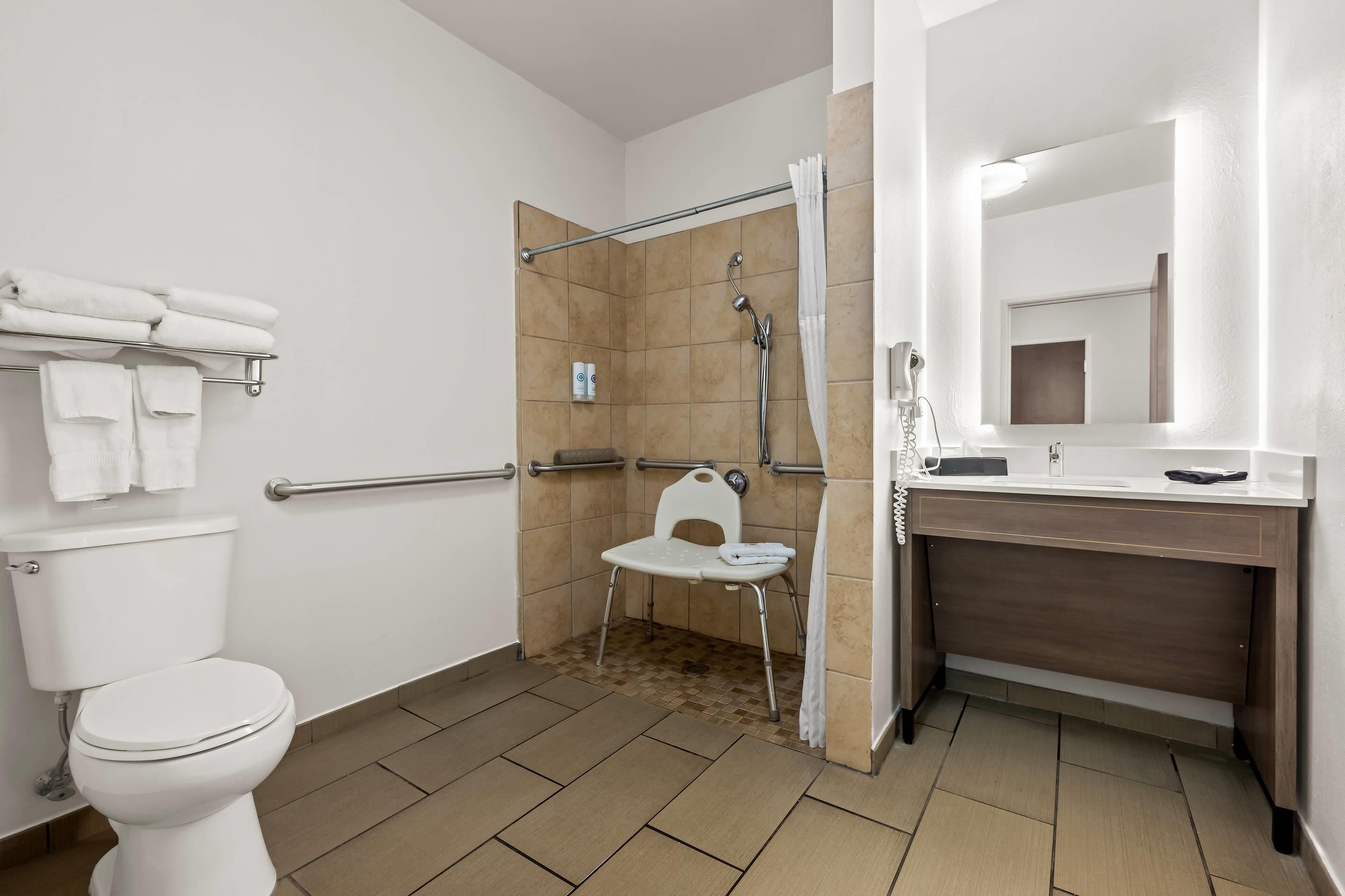 A bathroom with a toilet, a shower with a chair, a sink with a mirror, and a towel rack.