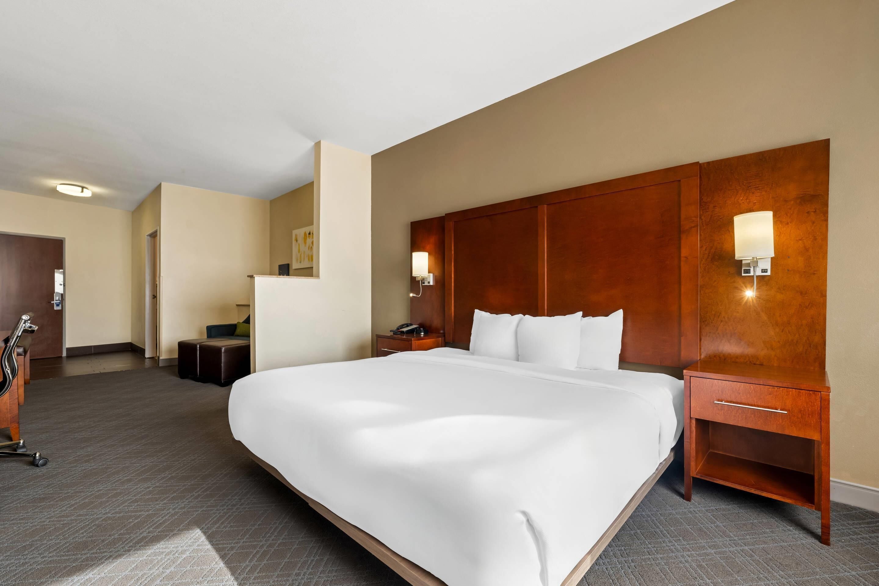 A hotel room with a large bed, wooden headboard, bedside tables with lamps, and a small seating area with a chair and ottoman.