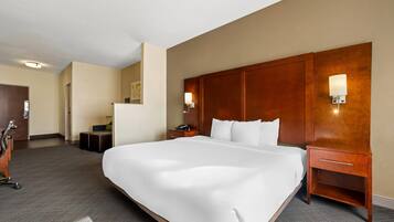 Quarto de hotel com cama grande, cabeceira de madeira, mesas de cabeceira com luminárias e um pequeno espaço de estar com poltrona e banquinho.