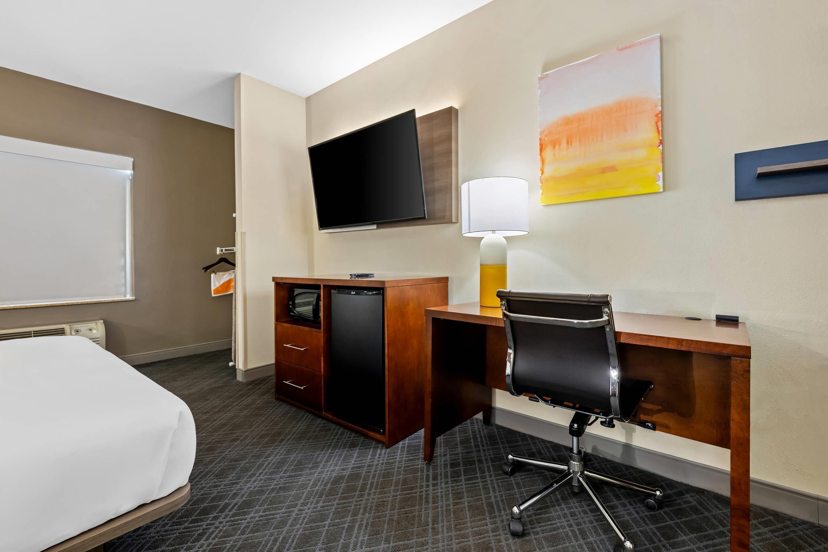 A hotel room with a bed, a desk with a chair, a television, a lamp, and a small cabinet.