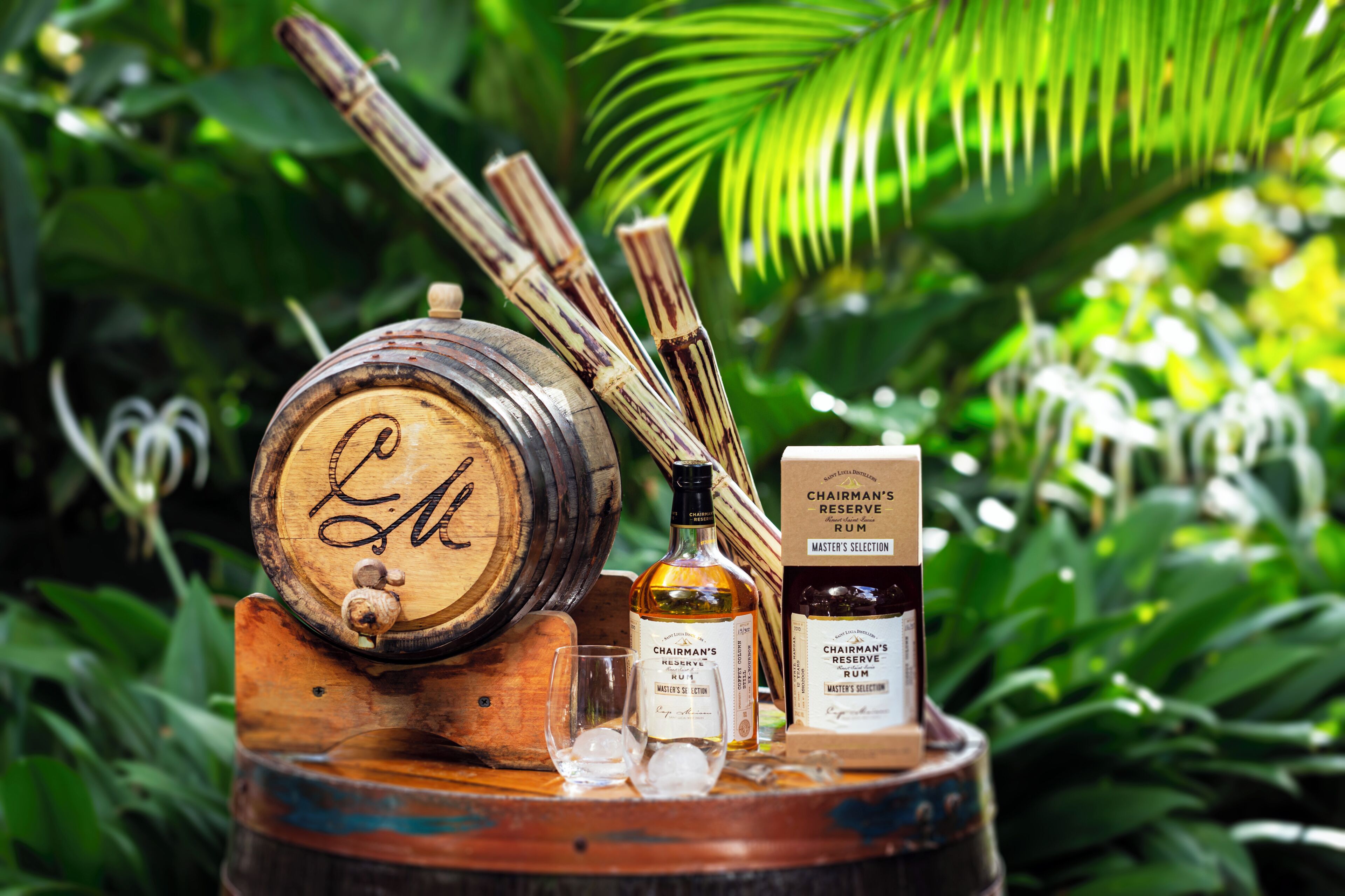 A wooden barrel with a label, a bottle of rum, and two glasses on a wooden stand surrounded by greenery.