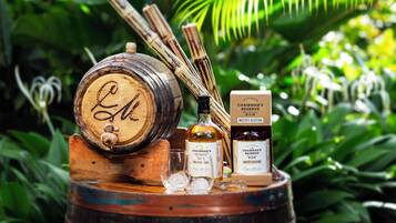 A wooden barrel with a label, a bottle of rum, and two glasses on a wooden stand surrounded by greenery.