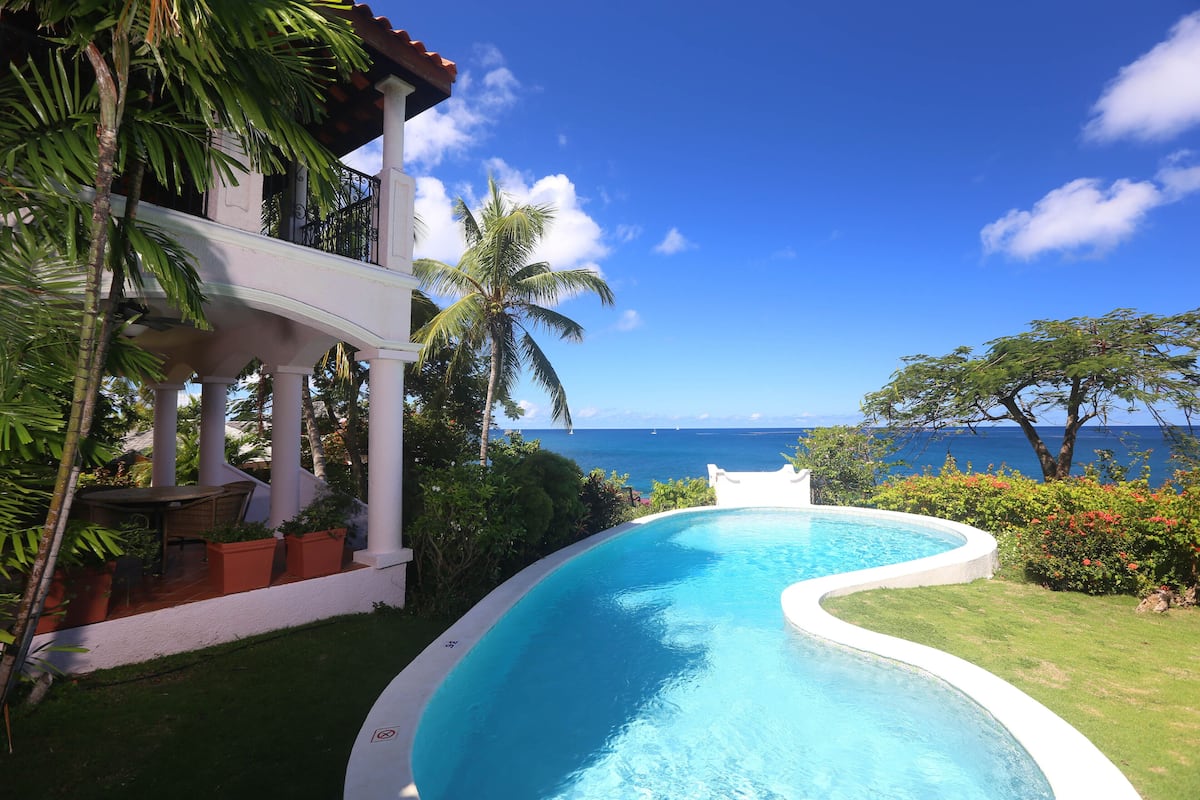 A curved pool with a view of the ocean, surrounded by lush greenery and a white building with a balcony.