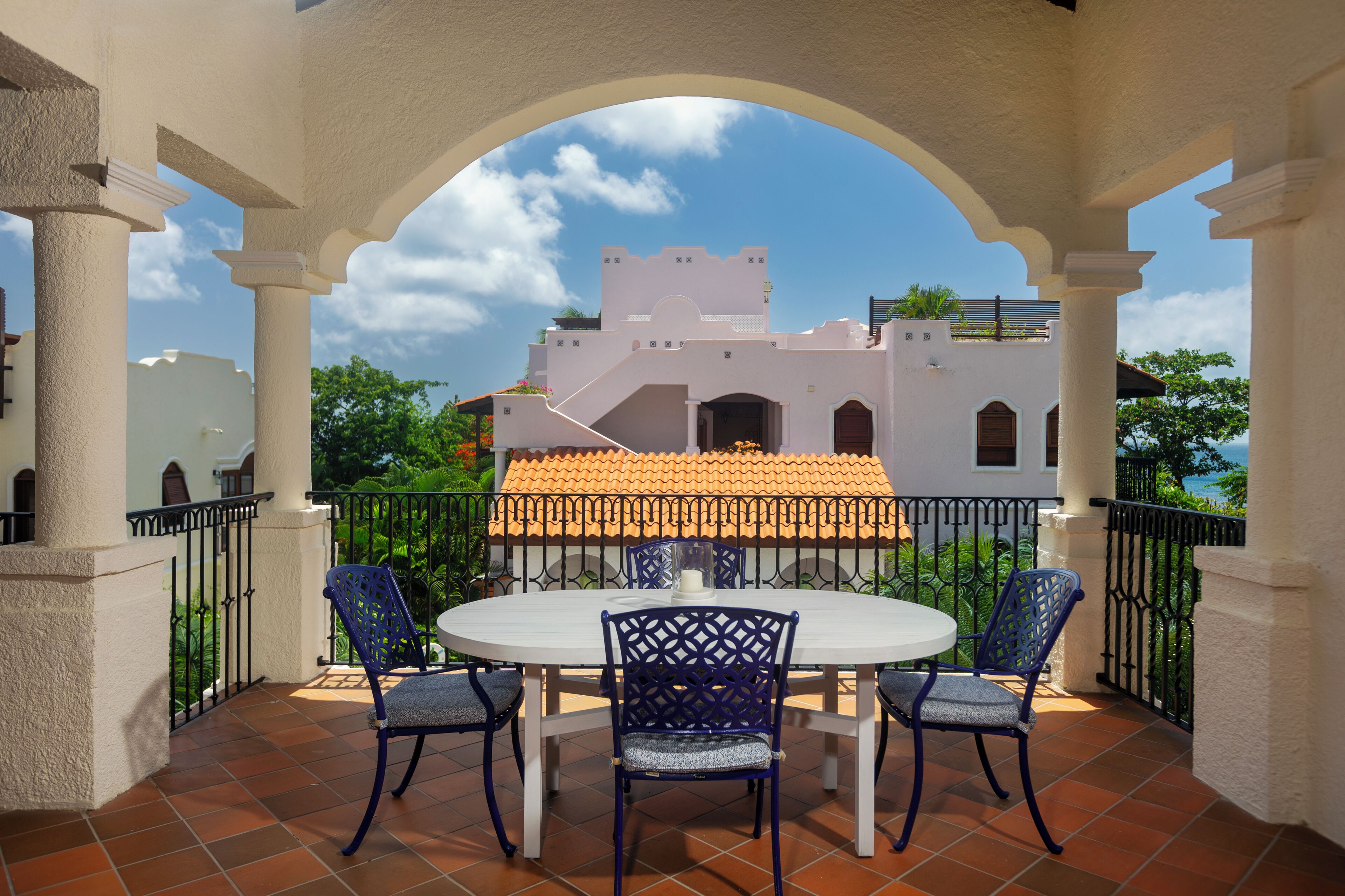 A patio with a round table and chairs, a view of a building with a tiled roof, and a clear sky.