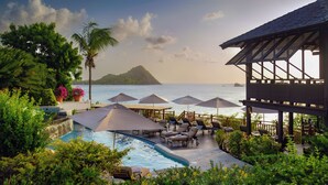 A pool area with lounge chairs and umbrellas, surrounded by lush greenery and overlooking a body of water with a distant island. - Cap Maison Resort & Spa (Gros Islet)