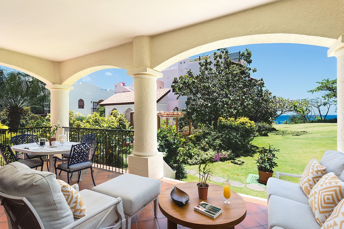A covered patio with outdoor seating, a table with a book and a glass of orange juice, and a view of a well-maintained garden and buildings in the background.