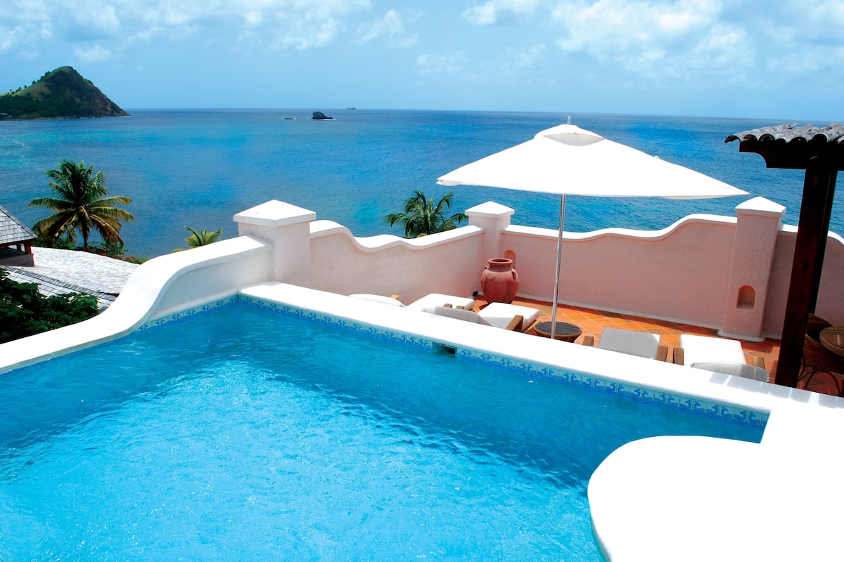 A pool with a view of the ocean, a white umbrella, and lounge chairs.