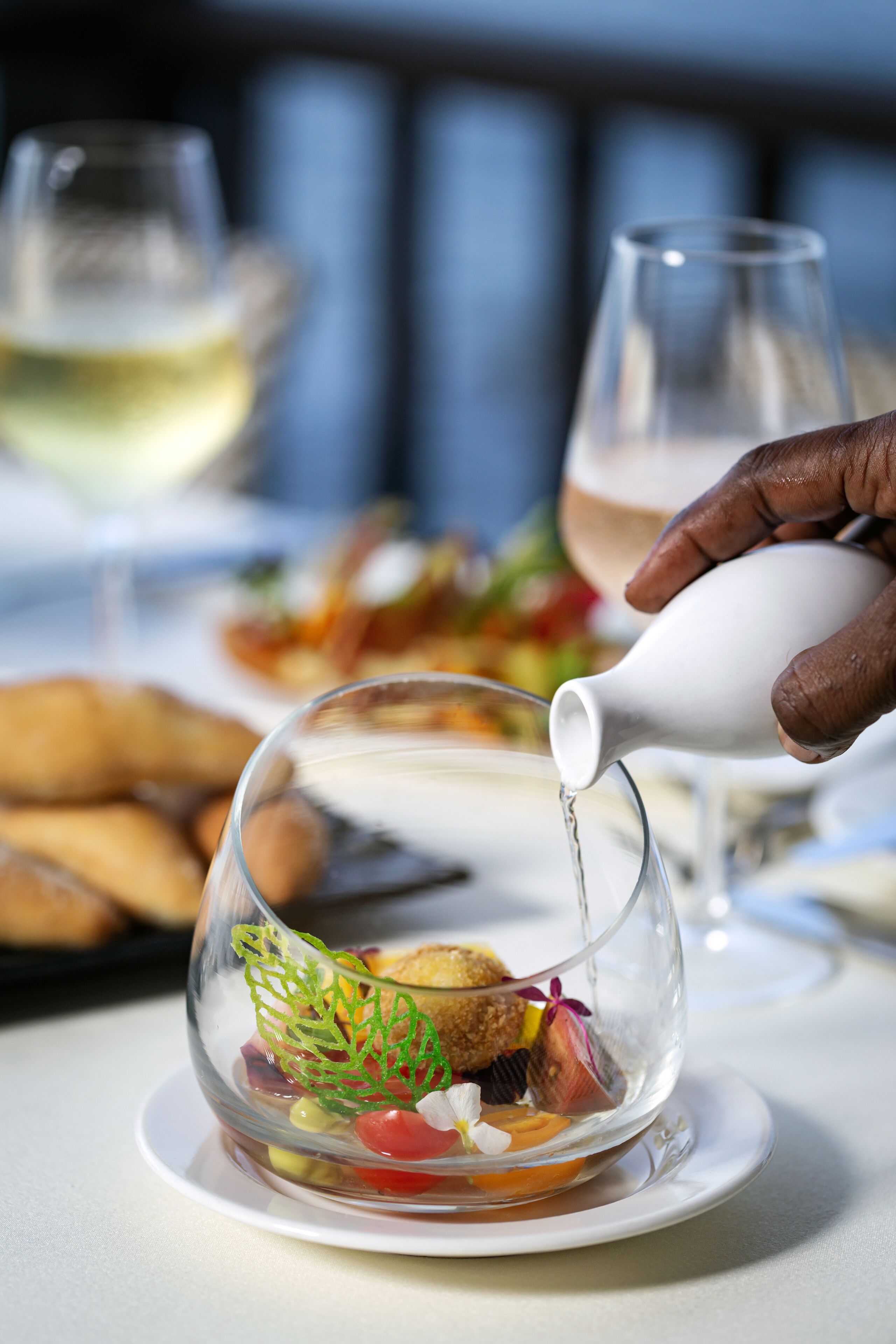 Ett matbord med en genomskinlig glaskupol som innehåller en dessert, en vit kanna som häller upp en dryck och ett glas vitt vin i bakgrunden.
