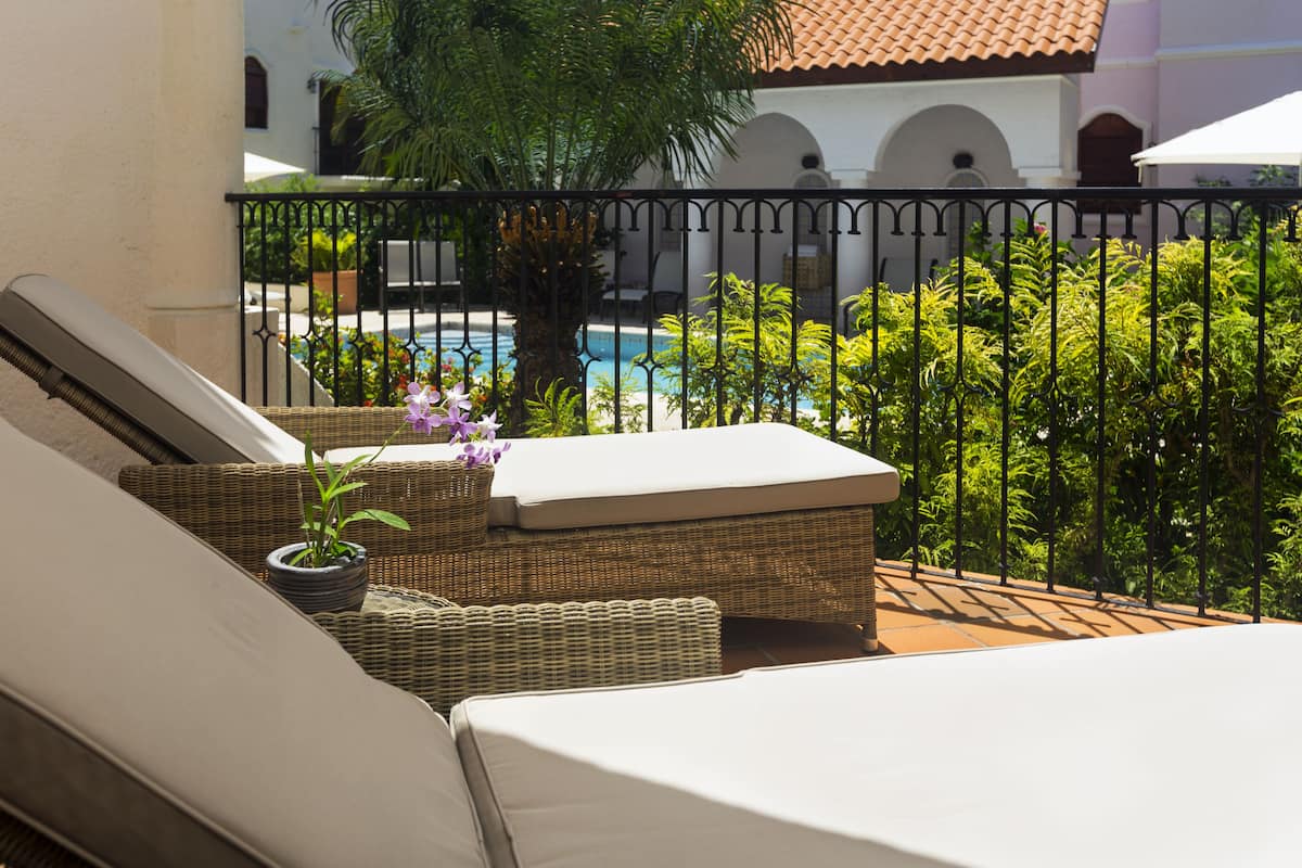 A balcony with wicker furniture, a potted plant, and a view of a swimming pool and greenery.