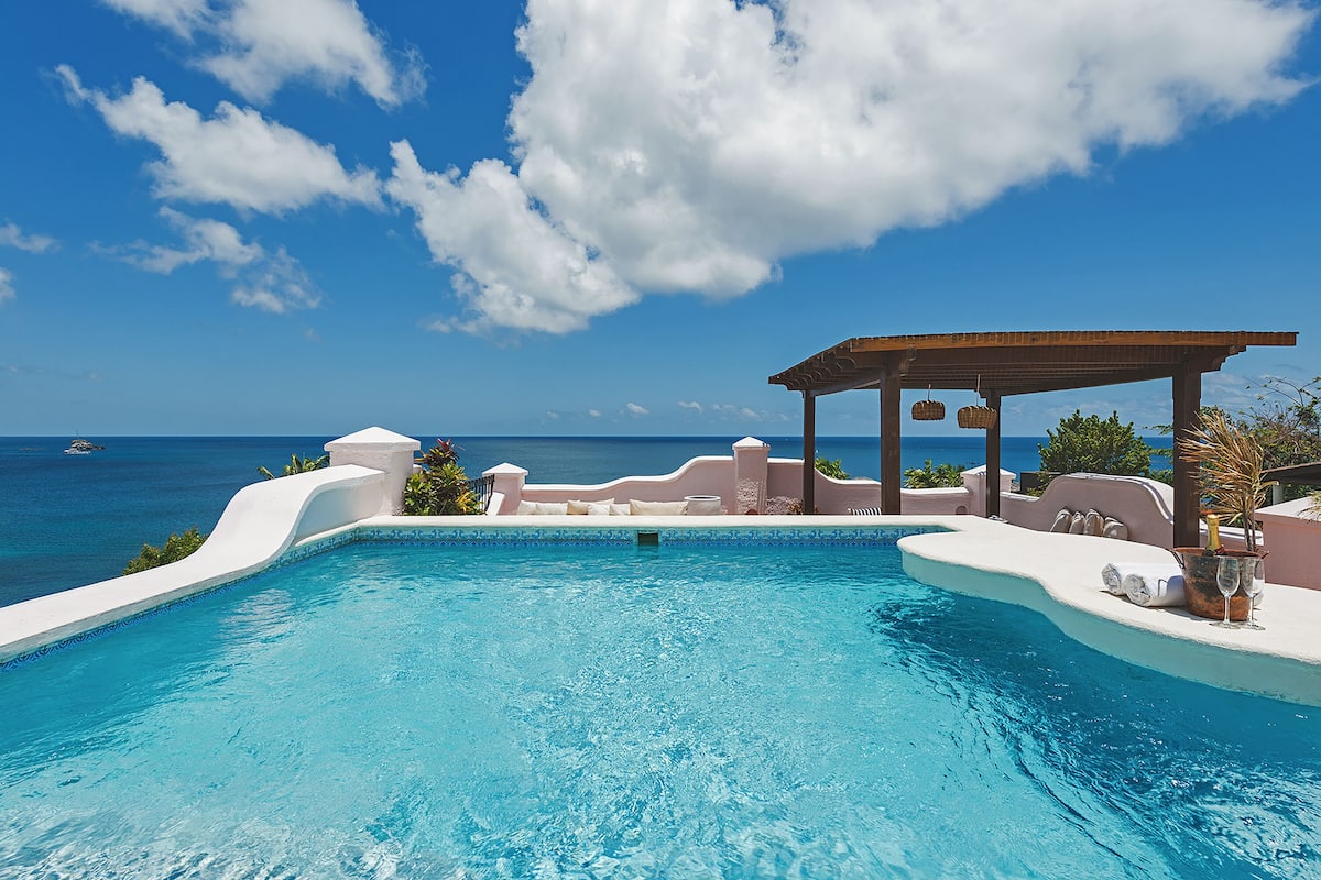 A pool with a view of the ocean, a pergola, and a lounge area with a drink.