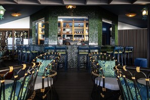A bar area with a green-tiled wall, wooden shelves, and blue chairs with patterned cushions.