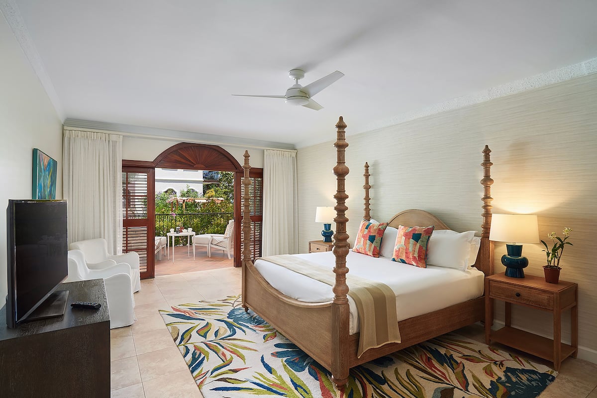 A four-poster bed with a television on a desk, a balcony with outdoor seating, and a ceiling fan.