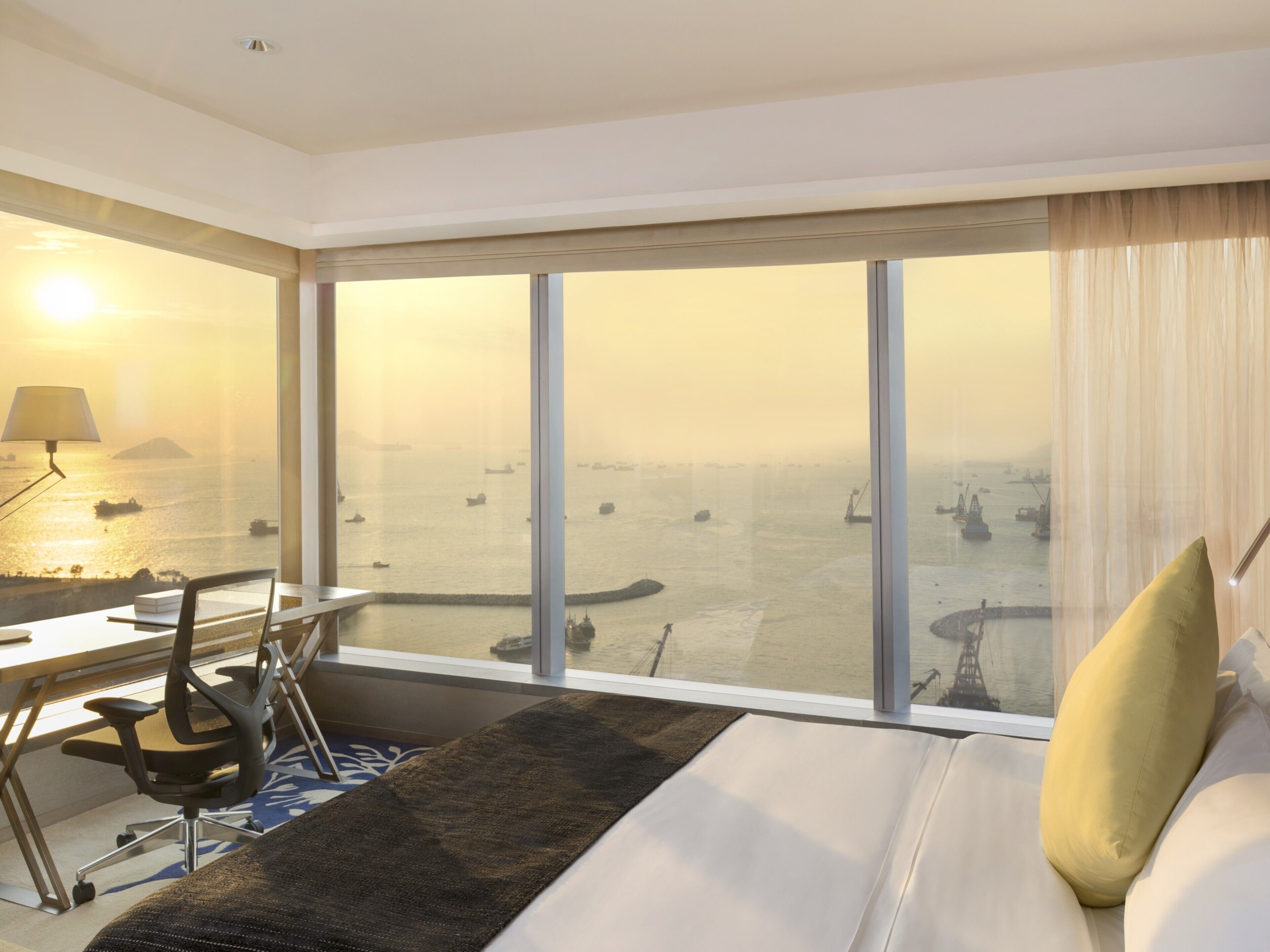 A hotel room with a large window, a desk with a chair, a bed, and a lamp.