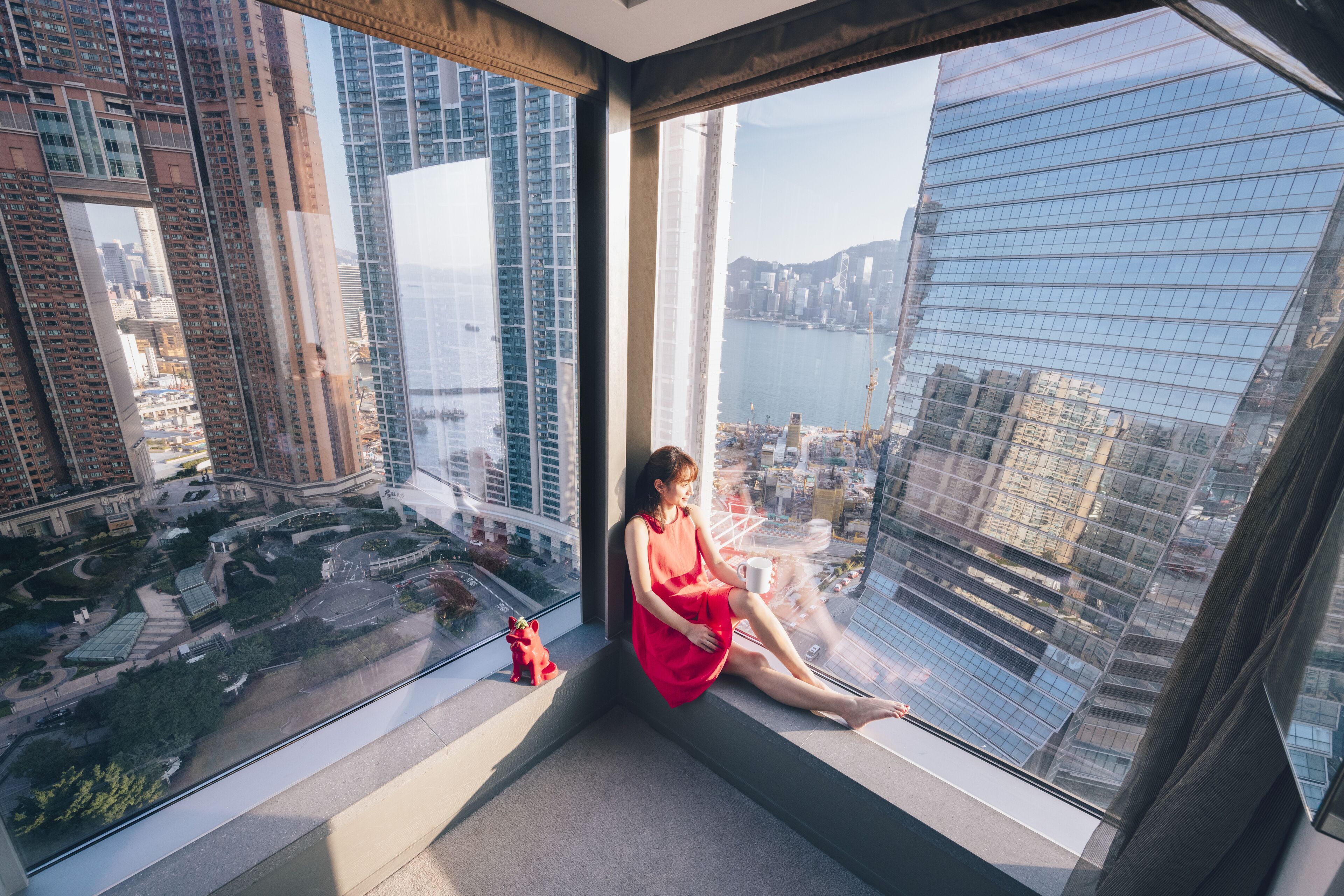 A woman sitting by a large window with a cityscape view, including high-rise buildings and a body of water.