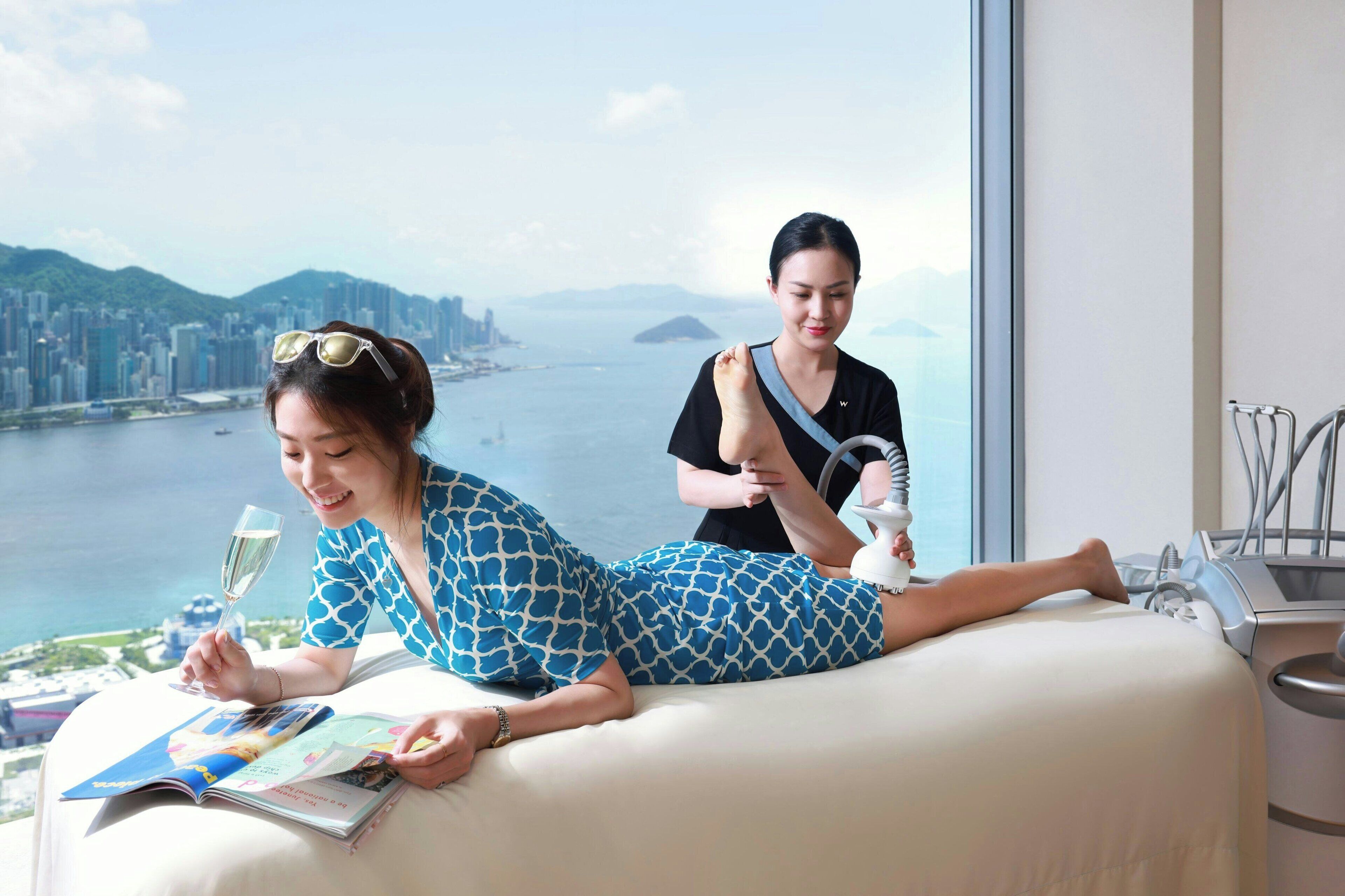 A woman lying on a bed reading a magazine with a cityscape view, another person applying a foot massage, and a window with a view of a body of water and mountains.