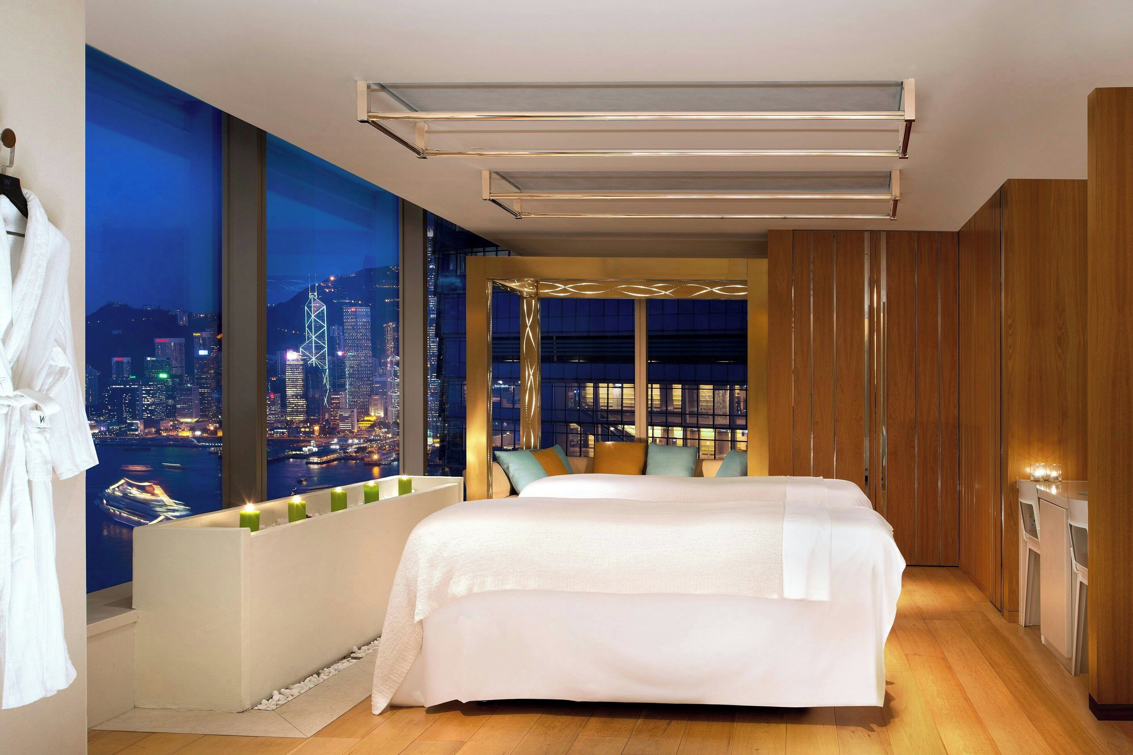 A modern hotel room with a large bed, a city view, a seating area with cushions, and a wooden wardrobe.