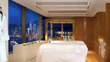 A modern hotel room with a large bed, a city view, a seating area with cushions, and a wooden wardrobe.
