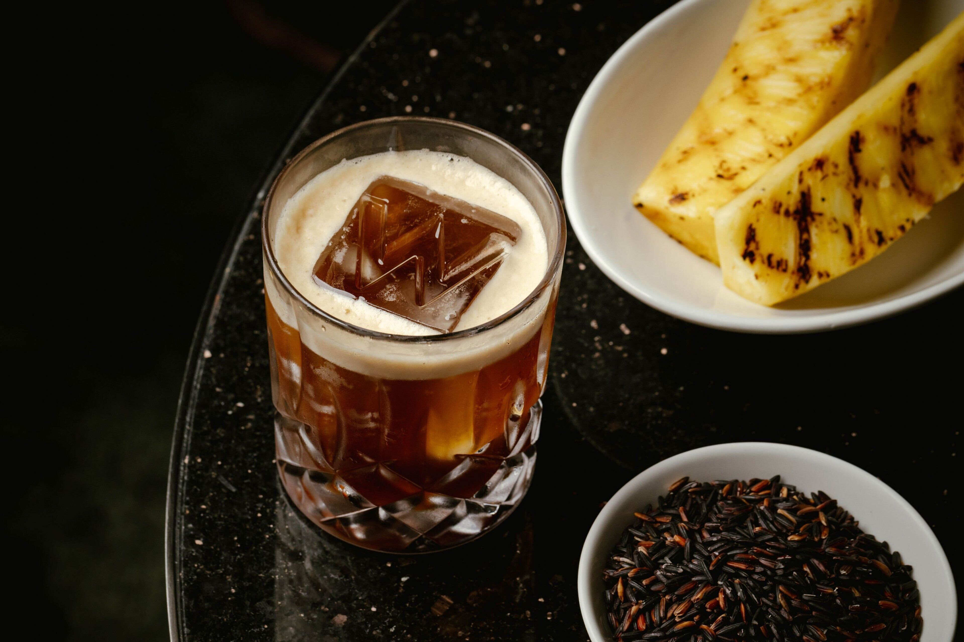 Un verre de thé glacé garni d’une tranche d’ananas sur le bord, un bol de riz noir et une assiette d’ananas grillé.