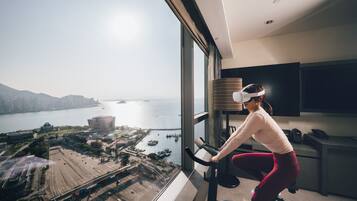 Uma pessoa usando uma bicicleta ergométrica em um quarto com uma grande janela com vista para uma paisagem urbana e para a água.