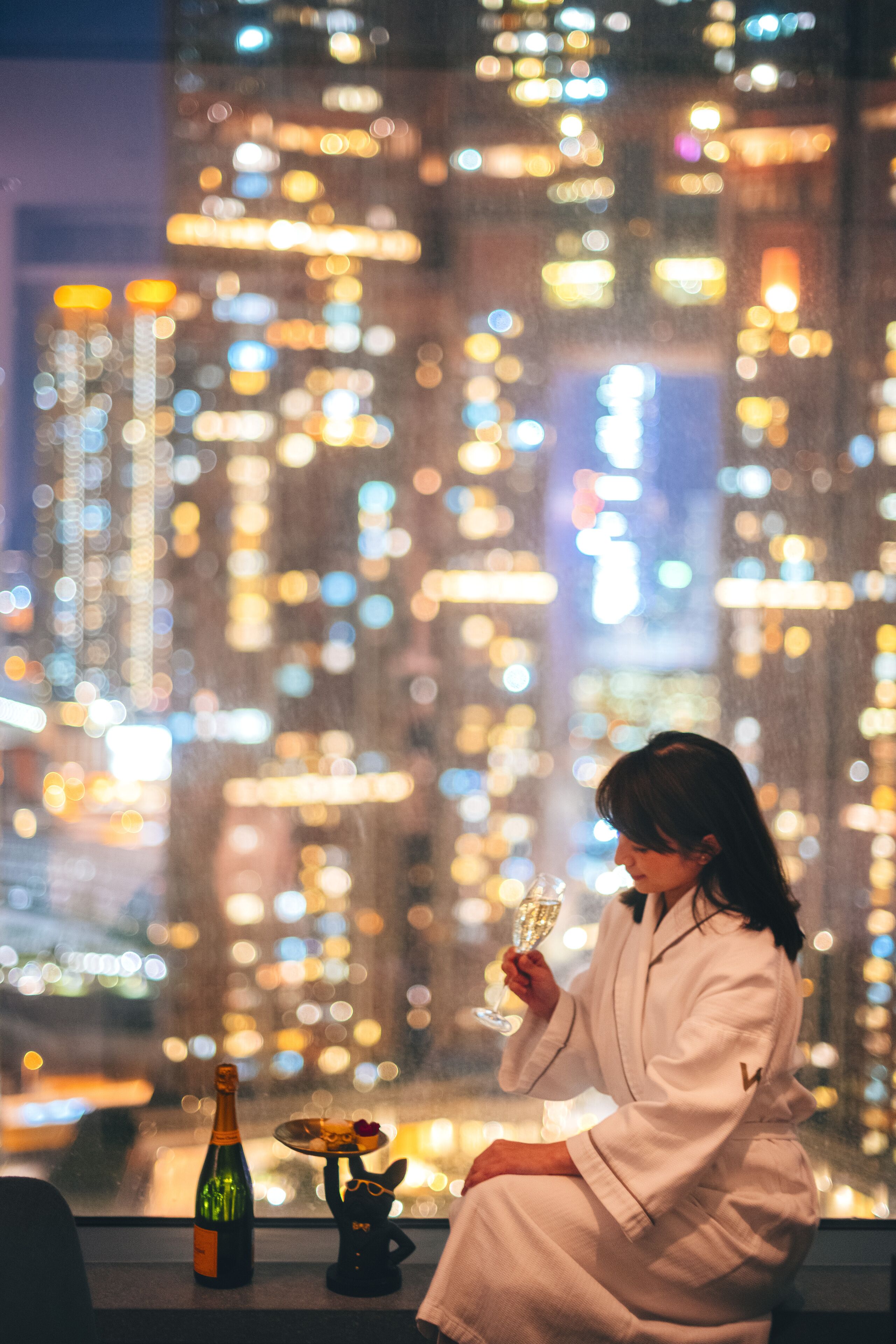 A person in a robe sitting by a window with a bottle of champagne and a plate of food.