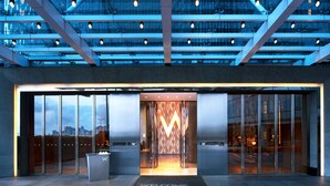 Une entrée d’hôtel moderne avec une façade en verre, un tapis d’accueil sur lequel est écrit « BIENVENUE » et une vue sur la ville.
