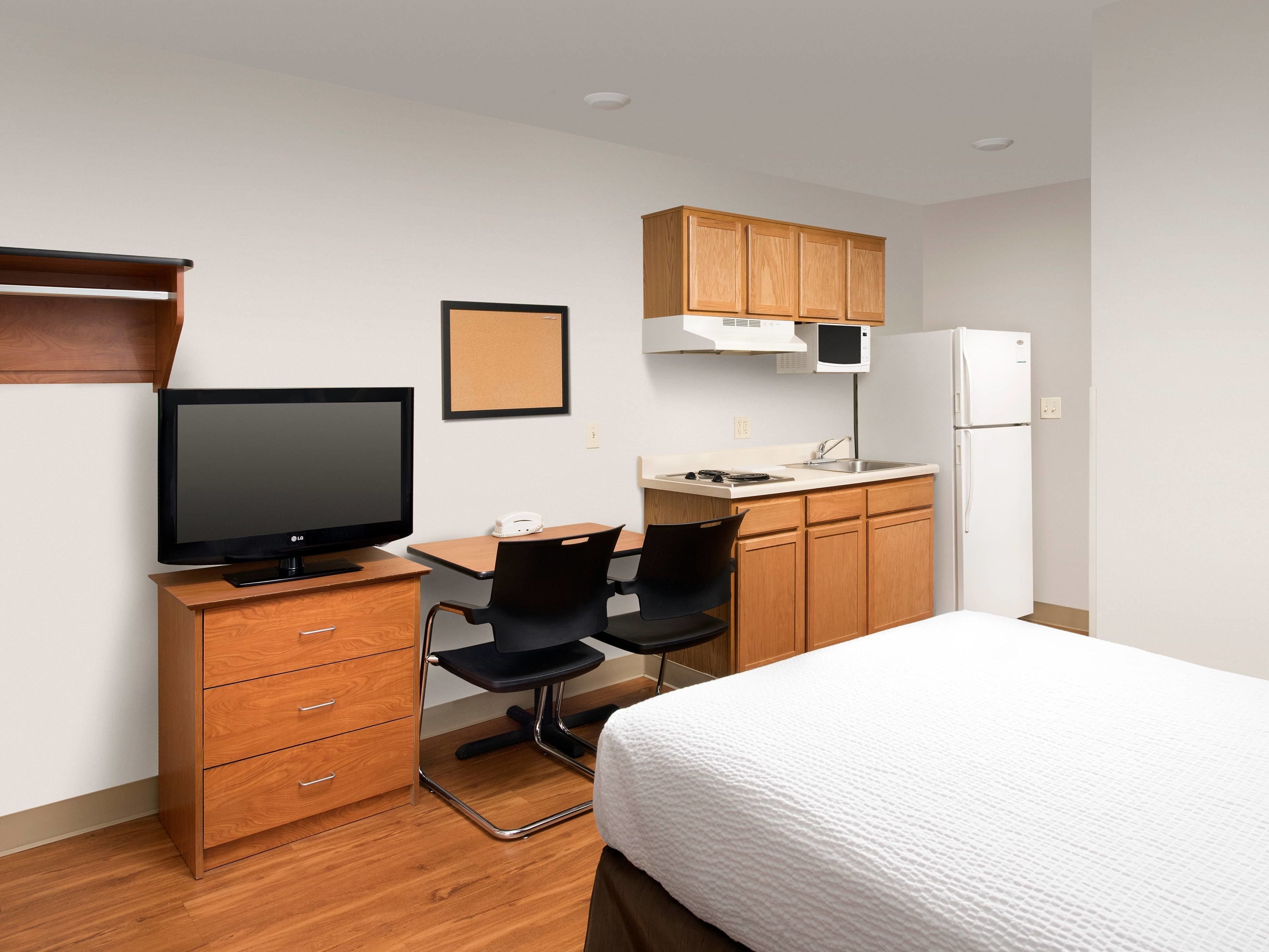 A compact hotel room with a bed, a desk with a computer, a chair, a small kitchenette, and a television.