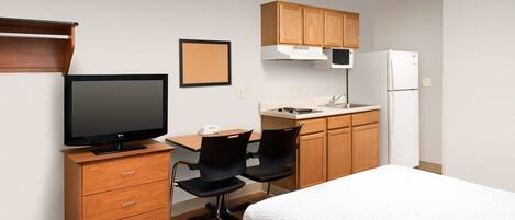 A compact hotel room with a bed, a desk with a computer, a chair, a small kitchenette, and a television.