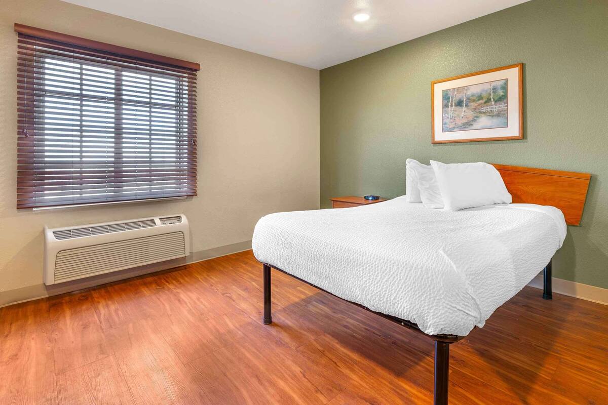 Un dormitorio con cama, mesita de noche, un cuadro en la pared y ventana con persianas.